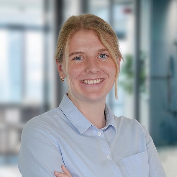 Eine Frau mit blonden Haaren in einem hellblauen Hemd lächelt in einem Büro mit unscharfem Hintergrund.