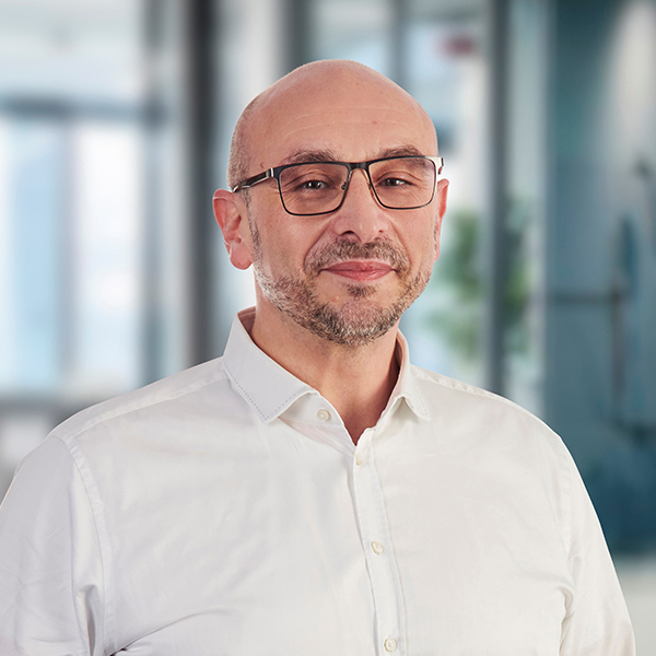 Ein kahlköpfiger Mann mit Brille, der ein weißes Hemd trägt, steht in einer modernen Büroumgebung und lächelt leicht.