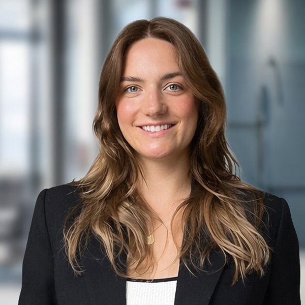 Eine Frau mit langen braunen Haaren, die einen schwarzen Blazer trägt, lächelt in einem Büro mit unscharfem Hintergrund.