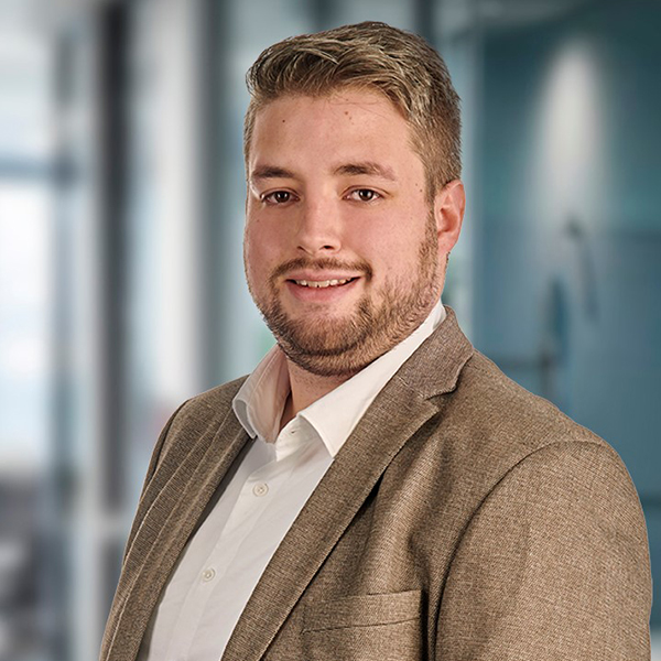 Ein Mann mit kurzen Haaren und Bart, der einen hellbraunen Blazer und ein weißes Hemd trägt und in einem Büro lächelt.