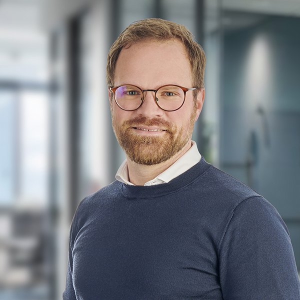 Ein Mann mit Brille und Bart, der einen blauen Pullover und ein weißes Hemd trägt und in einem Büro steht.