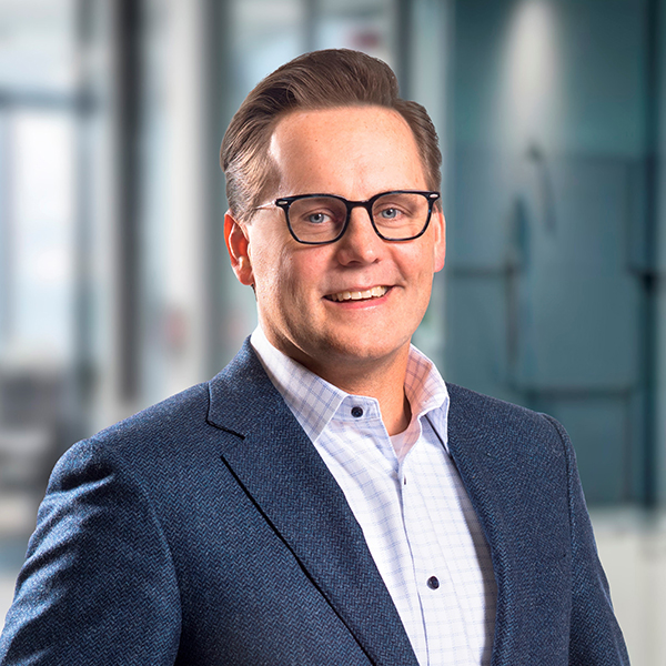Ein lächelnder Mann mit Brille, einem blauen Blazer und einem weiß karierten Hemd in einer modernen Büroumgebung.