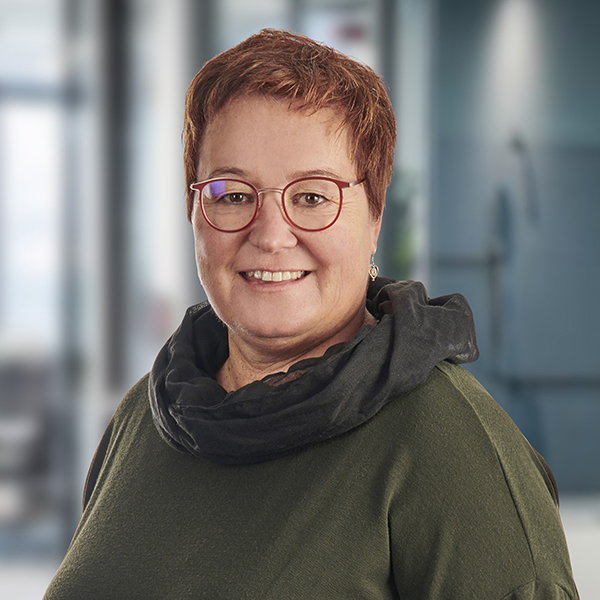 Lächelnde Frau mit kurzen Haaren, roter Brille und dunklem Schal, die in einer modernen Büroumgebung steht.