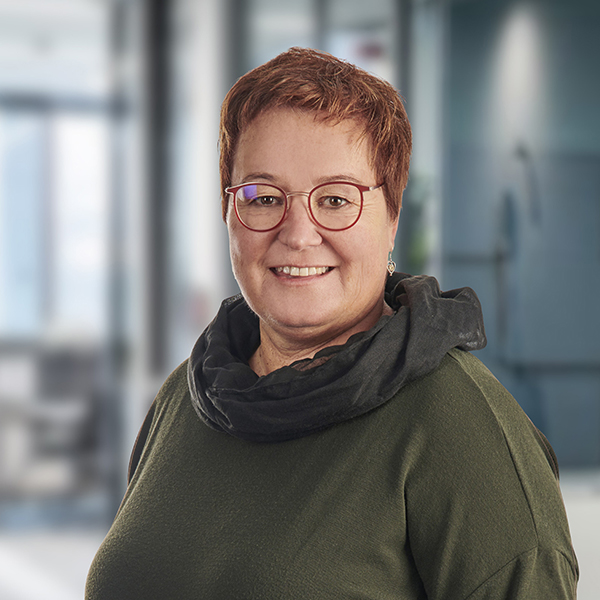 Eine lächelnde Person mit kurzen Haaren, Brille und einem dunklen Schal, die in einer modernen, unscharfen Büroumgebung steht.
