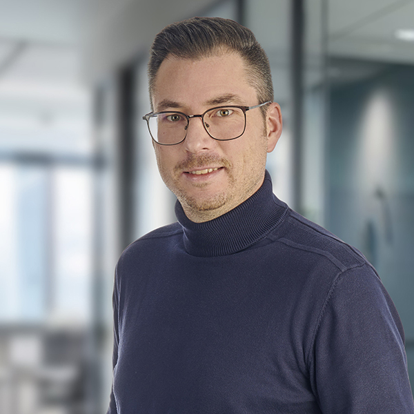 Ein Mann mit Brille in einem marineblauen Rollkragenpullover, der in einem modernen Büro mit unscharfem Hintergrund steht.