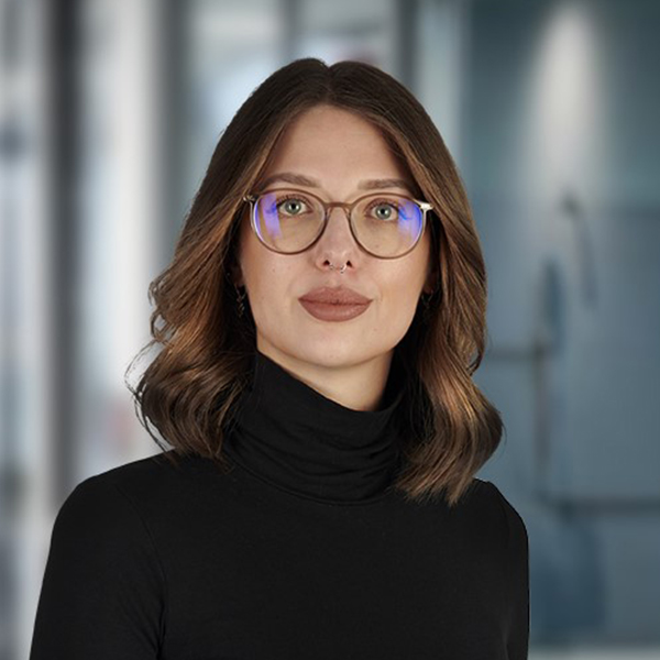 Eine Frau mit braunen Haaren, Brille und einem schwarzen Rollkragenpullover, die in einer unscharfen Büroumgebung steht.
