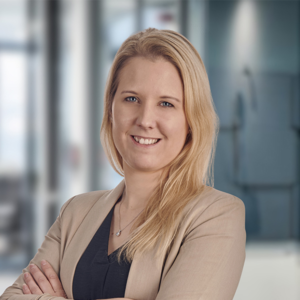 Eine Frau mit langen blonden Haaren in einem beigen Blazer, die in einer modernen Büroumgebung lächelt.