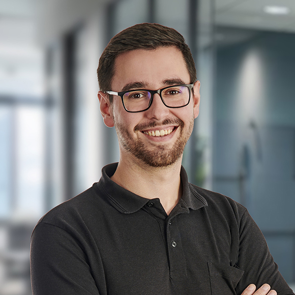 Ein lächelnder Mann mit Brille und Bart steht in einem Büroflur und trägt ein schwarzes Poloshirt.