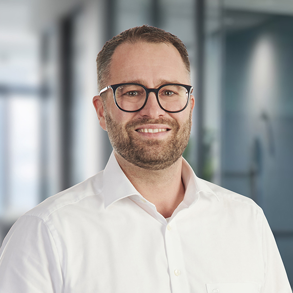 Ein lächelnder Mann mit Brille und Bart, der ein weißes Hemd trägt, steht in einem Büroflur.