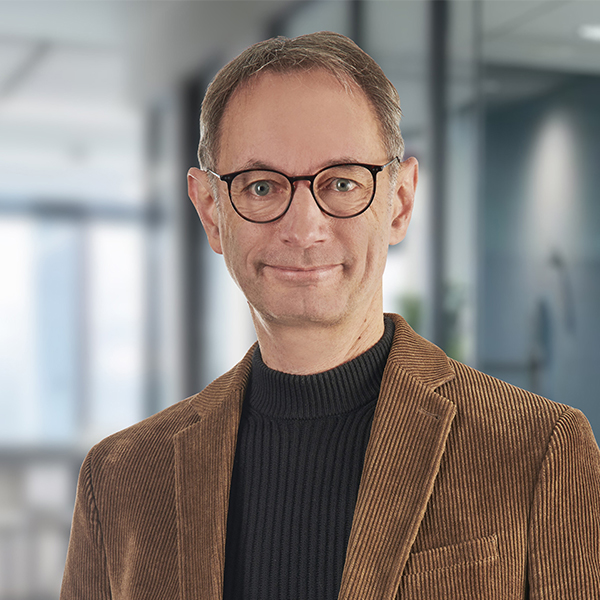 Ein lächelnder Mann mit Brille und braunem Blazer in einer Büroumgebung, der in die Kamera blickt.