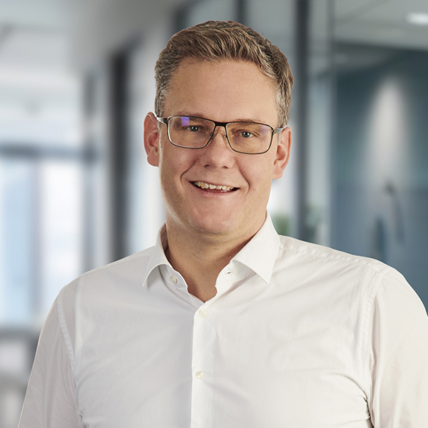 Ein lächelnder Mann mit Brille, der ein weißes Hemd trägt und in einem modernen Büro mit unscharfem Hintergrund steht.