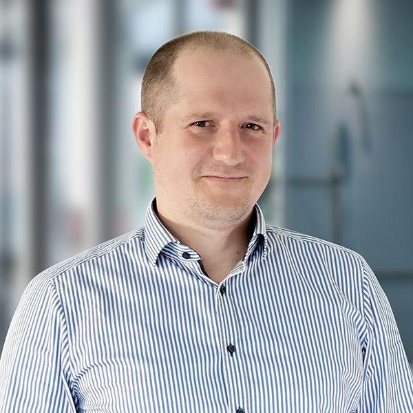 Ein glatzköpfiger Mann in einem gestreiften Hemd, der lächelt und vor einem unscharfen Bürohintergrund steht.