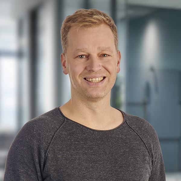 Ein lächelnder Mann mit kurzen blonden Haaren und einem grauen Hemd steht in einer modernen, unscharf abgebildeten Büroumgebung.