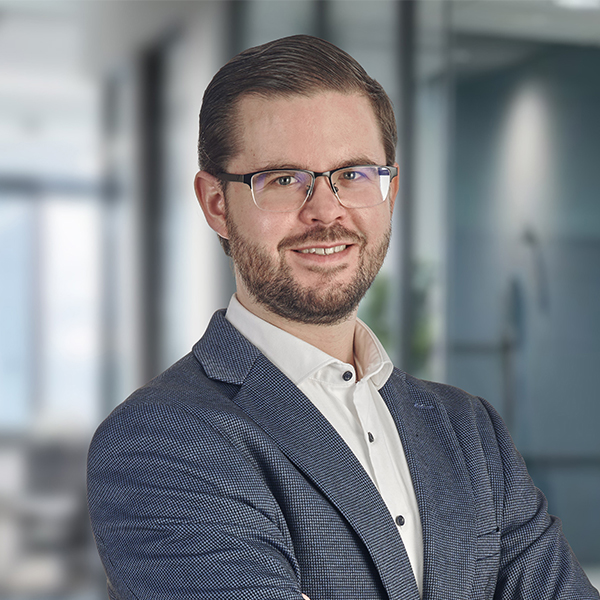Ein Mann im Anzug und mit Brille lächelt mit verschränkten Armen in einer modernen Büroumgebung.